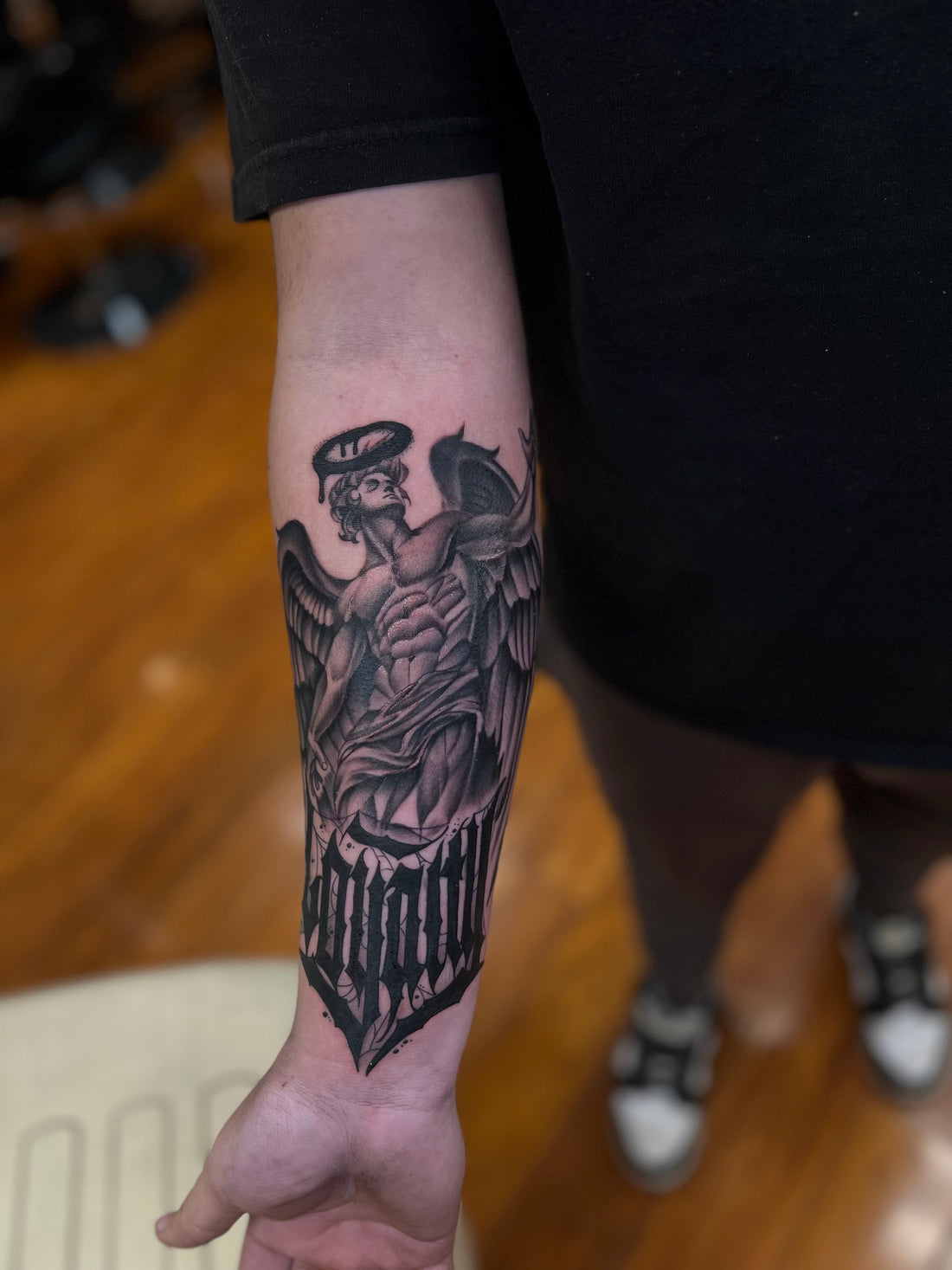 Black and grey archangel forearm tattoo with heavy black lettering at the wrist, tattooed at Tagz Tattoo in Mesa, AZ