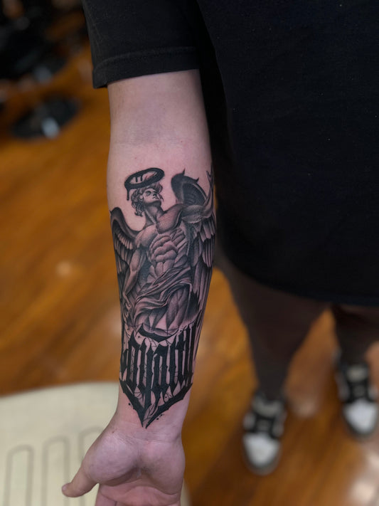 Black and grey archangel forearm tattoo with heavy black lettering at the wrist, tattooed at Tagz Tattoo in Mesa, AZ